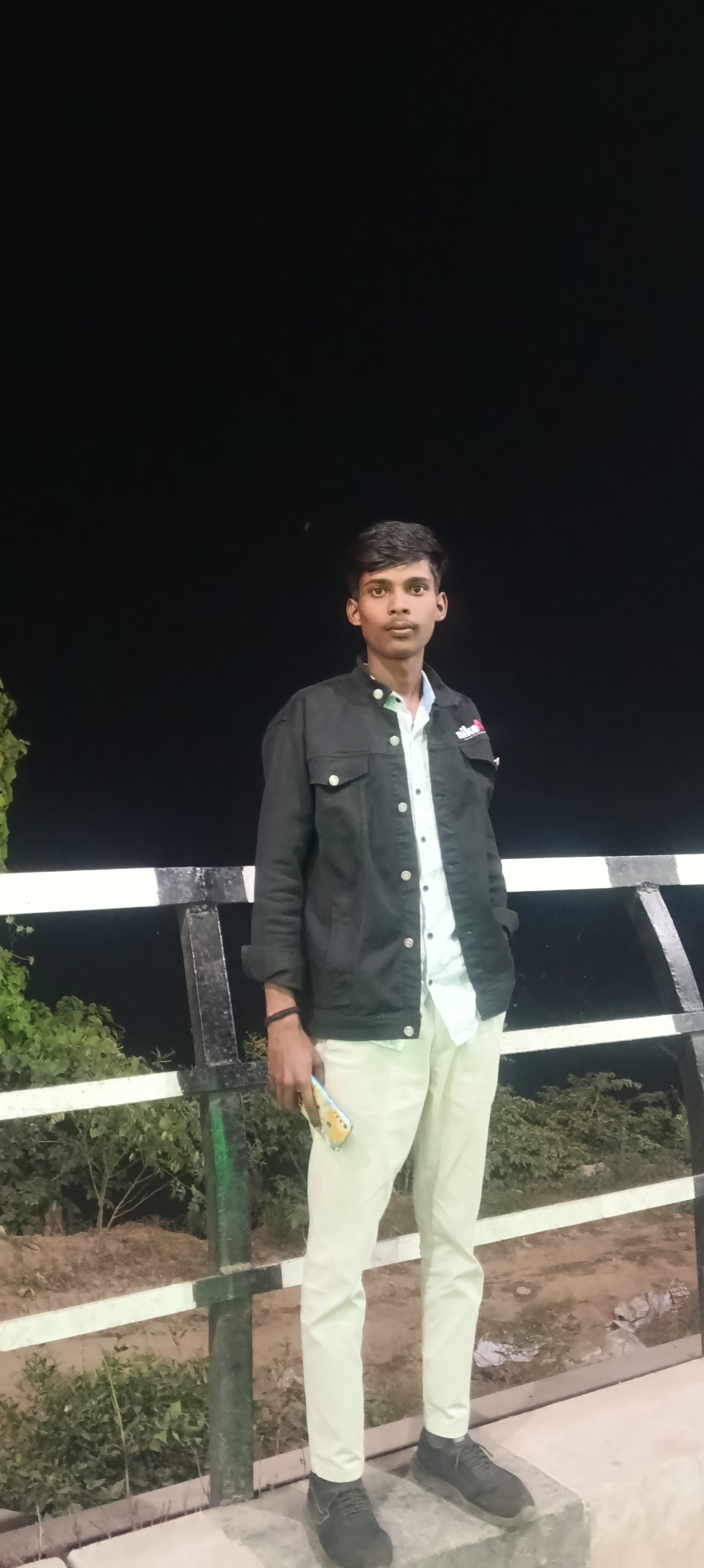 young man walks alone on a dimly lit path at night, wearing dark casual clothes and white sneakers. Beside him appears a Golden glowing, Deon-style illustration of a girl holding a bouquet of bright red roses. The neon girl emits a soft white light, contrasting against the dark, blurry background of city lights in the distance. The atmosphere feels calm, melancholic, and slightly surreal, blending reality and imagination. 100% accurate my face, both holding hand middle of road - AI Generated Image